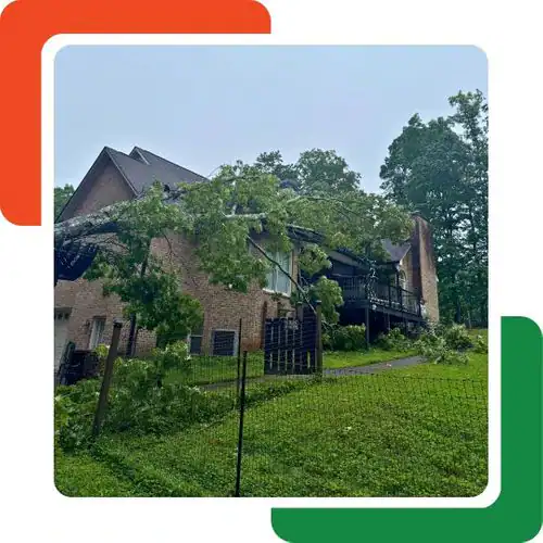 A large tree has fallen onto the roof of a brick house on a grassy hill in Cherokee, GA, causing visible damage. The sky is clear, and there is a black metal fence in the foreground—tree removal Cherokee services may be needed.